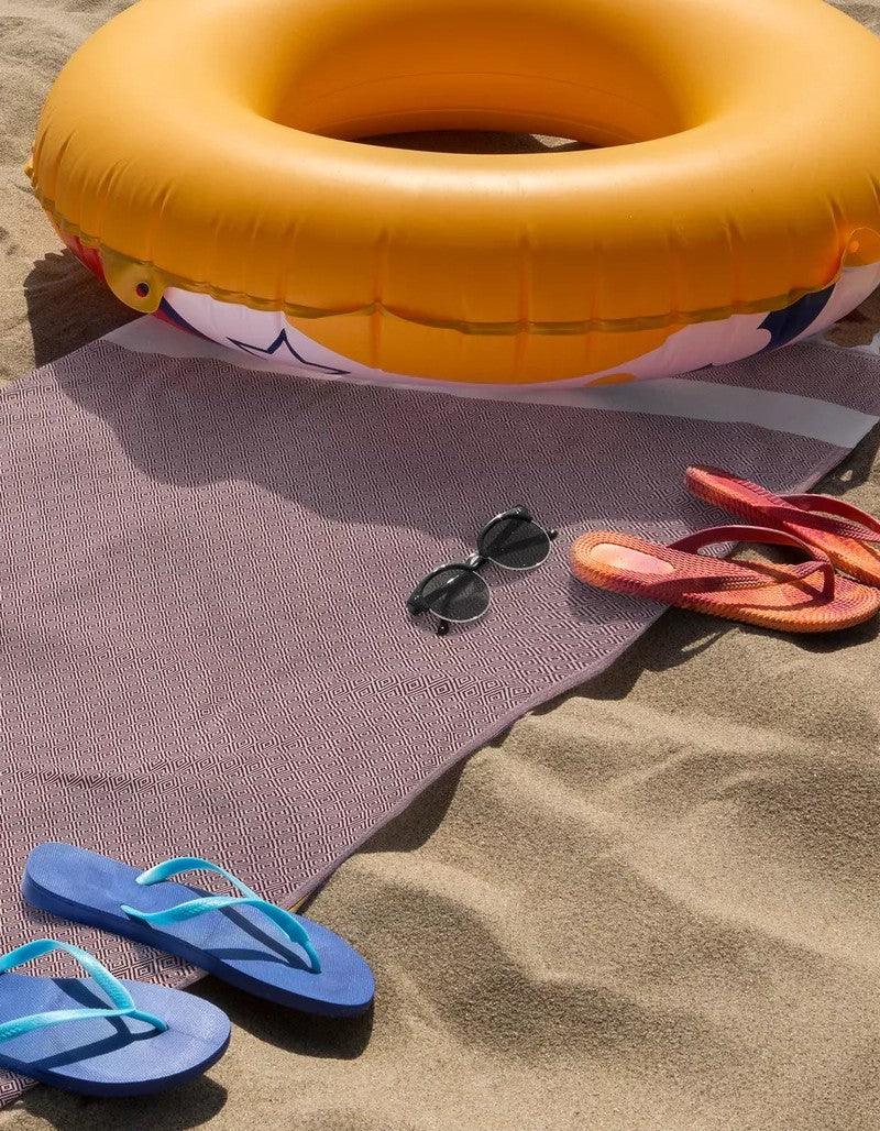 Waschbarer Strandteppich mit geometrischem Muster, Sonnenbrille und Flipflops auf Sand neben aufblasbarem Ring